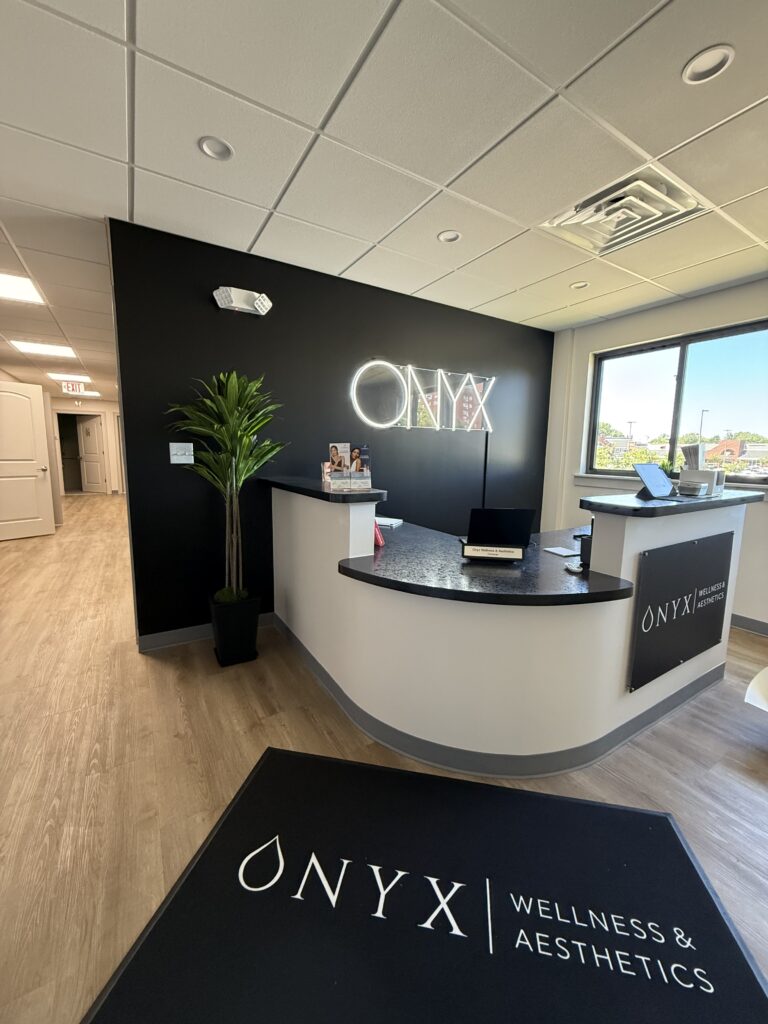 Modern reception area with a black accent wall featuring a neon ONYX sign, a reception desk with a laptop, and a welcome mat that reads "ONYX WELLNESS & AESTHETICS."