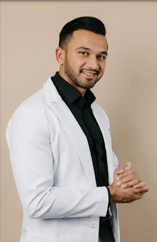A smiling man in a white lab coat over a black shirt, standing with hands clasped against a beige background.